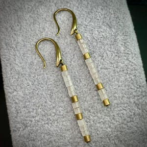 May include: A pair of long, gold earrings with white beads. The earrings have a simple, elegant design.