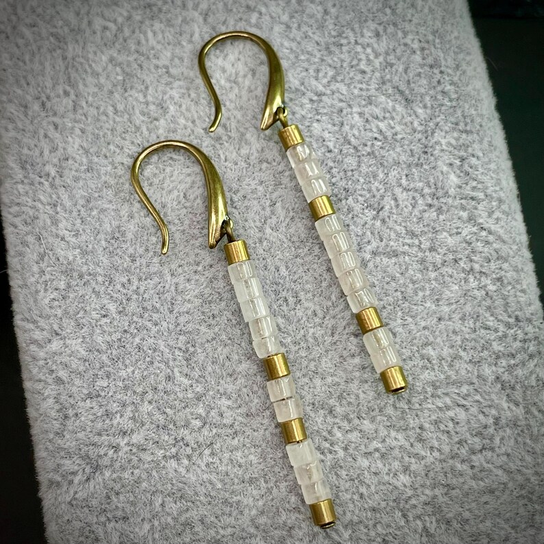May include: A pair of long, gold earrings with white beads. The earrings have a simple, elegant design.