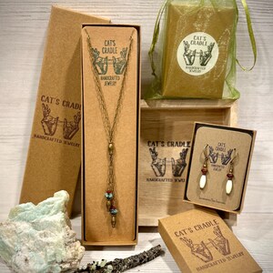 May include: Three brown cardboard boxes with the text "Cat's Cradle Handcrafted Jewelry" on them. The boxes contain a necklace, earrings, and a small bag. The necklace is made of gold chain with turquoise beads and a gold pendant. The earrings are made of gold with turquoise and white beads. The bag is made of green mesh.