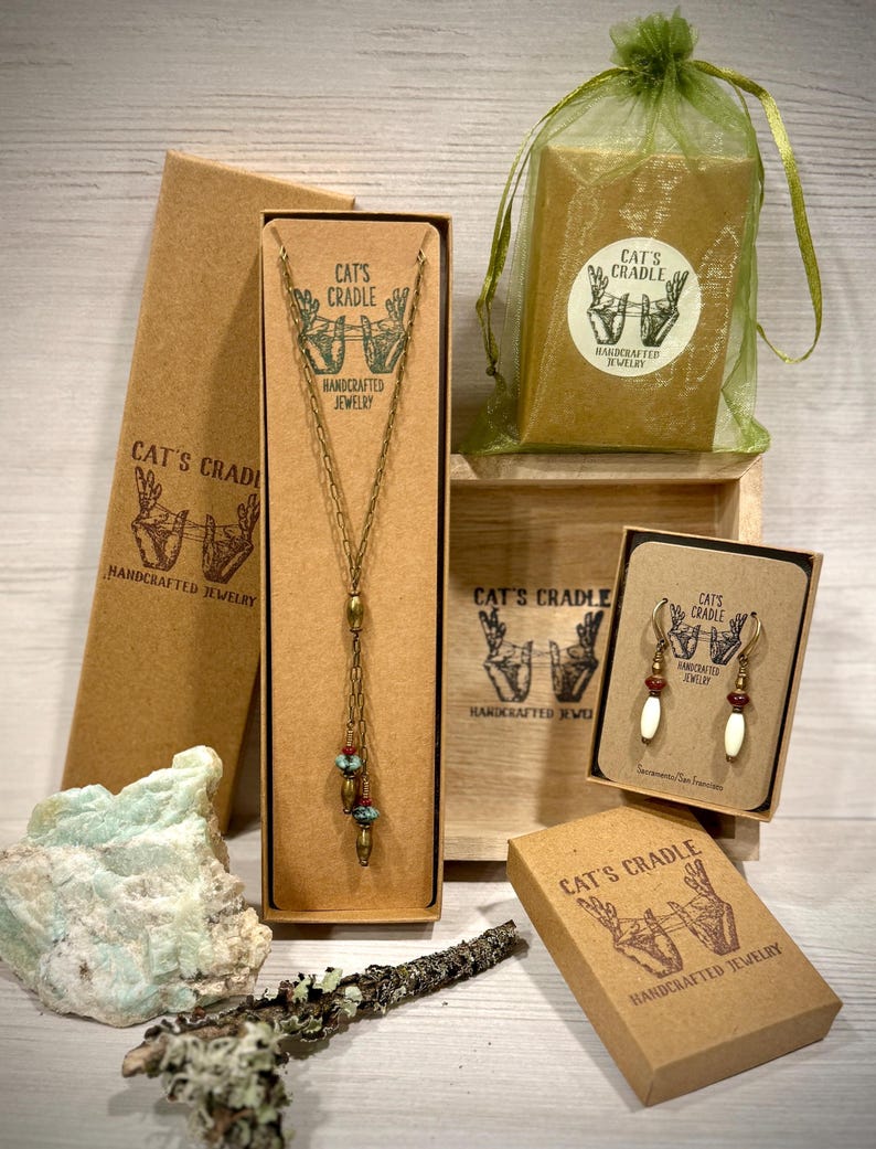 May include: Three brown cardboard boxes with the text "Cat's Cradle Handcrafted Jewelry" on them. The boxes contain a necklace, earrings, and a small bag. The necklace is made of gold chain with turquoise beads and a gold pendant. The earrings are made of gold with turquoise and white beads. The bag is made of green mesh.
