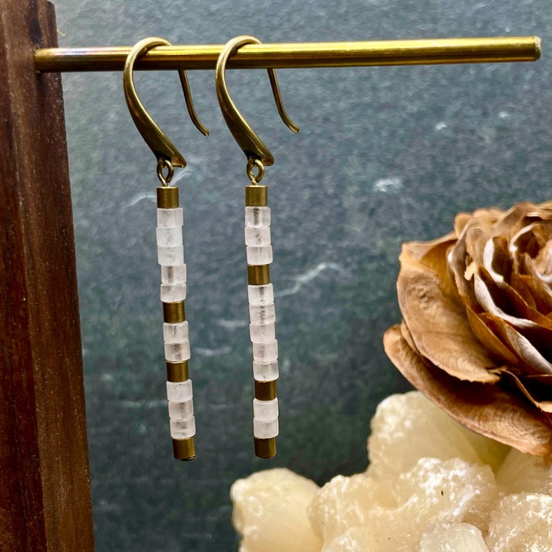May include: A pair of gold earrings with white, square-shaped beads hanging from the bottom. The earrings are hanging from a gold stand.