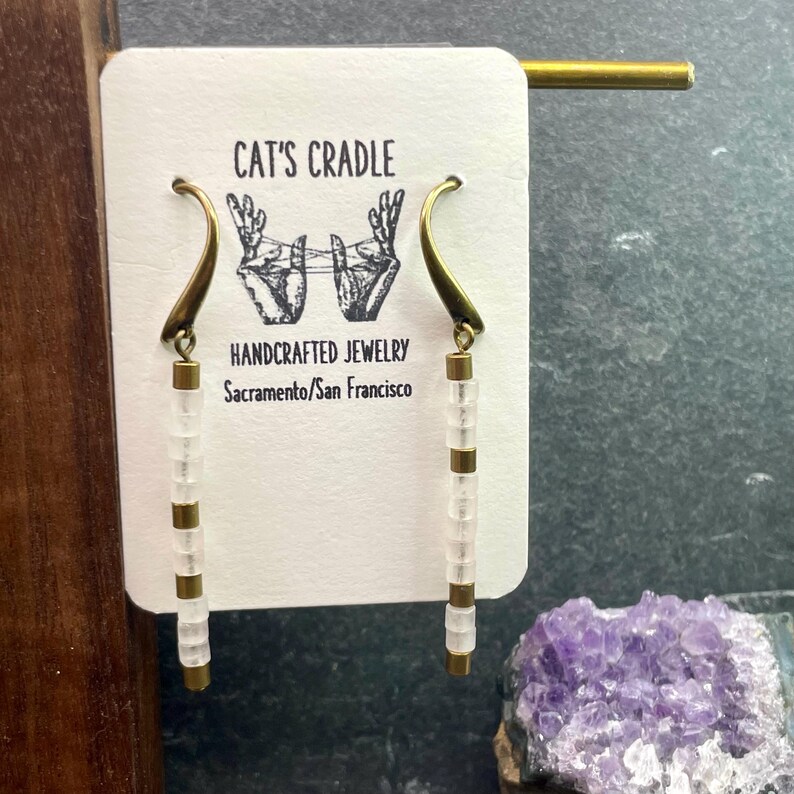 May include: A pair of white bead earrings with brass hooks. The earrings are hanging from a card that says "Cat's Cradle Handcrafted Jewelry Sacramento/San Francisco".