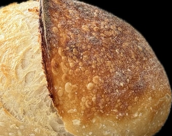 May include: A round loaf of sourdough bread with a deeply scored top. The crust features a gradient of colors, transitioning from a light, pale golden-yellow on the left side to a dark, toasted brown on the right side, with a visible texture.