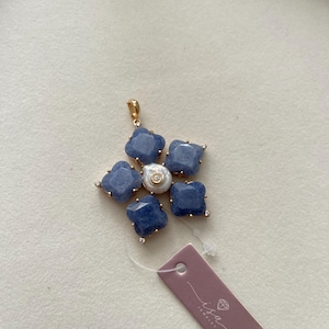 May include: A gold-toned pendant in a flower shape, featuring six blue-hued, square-cut gemstones surrounding a central pearl. Small, clear stones accent the edges. A tag with the "isa jewelry" logo is attached.