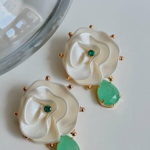 May include: A pair of white flower-shaped earrings with green gemstone accents. Each earring features a layered flower design with a small green gemstone in the center and a larger teardrop-shaped green gemstone dangling below.