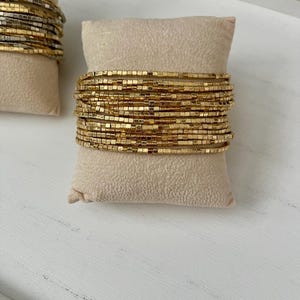 May include: Gold beaded bracelets displayed on a beige pillow. The bracelets are made of small, square gold beads, stacked in multiple rows. The bracelets are a shiny gold color and are presented on a neutral background.