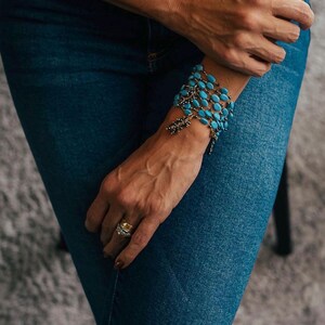 May include: A turquoise blue beaded bracelet with a silver charm and a gold ring on a person's finger.