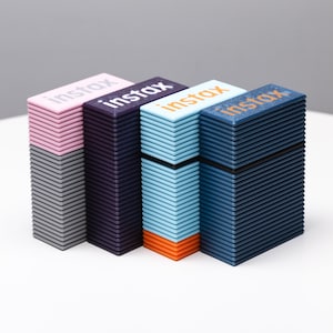 May include: Four stacks of instant film in different colors: gray, purple, blue, and dark blue. Each stack has the word "instax" printed on the top.