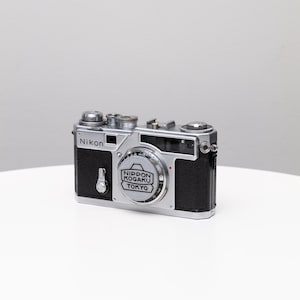 May include: A vintage Nikon camera with a silver body and black leather grip. The camera has a lens with the text "NIPPON KOGAKU TOKYO" and the Nikon logo. The camera is on a white surface.