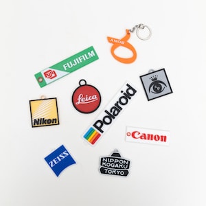 Op de afbeelding: Een verzameling sleutelhangers en labels met logo's van cameramerken, waaronder Fujifilm, Leica, Polaroid, Canon, Nikon, Zeiss en Nippon Kogaku Tokyo. De sleutelhangers zijn in verschillende vormen en kleuren, met een Sony sleutelhanger in oranje.