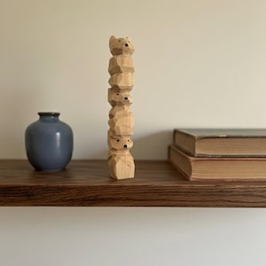 Puede incluir: Una escultura de madera de osos apilados, tallada en madera de color claro. Los osos están estilizados con rasgos simples y una textura tosca. Un jarrón azul y dos libros están en una estantería de madera.