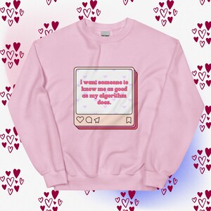 May include: A pale pink sweatshirt featuring the phrase "i want someone to know me as good as my algorithm does." The text is within a pink and white social media post design. The background has a pattern of hearts.