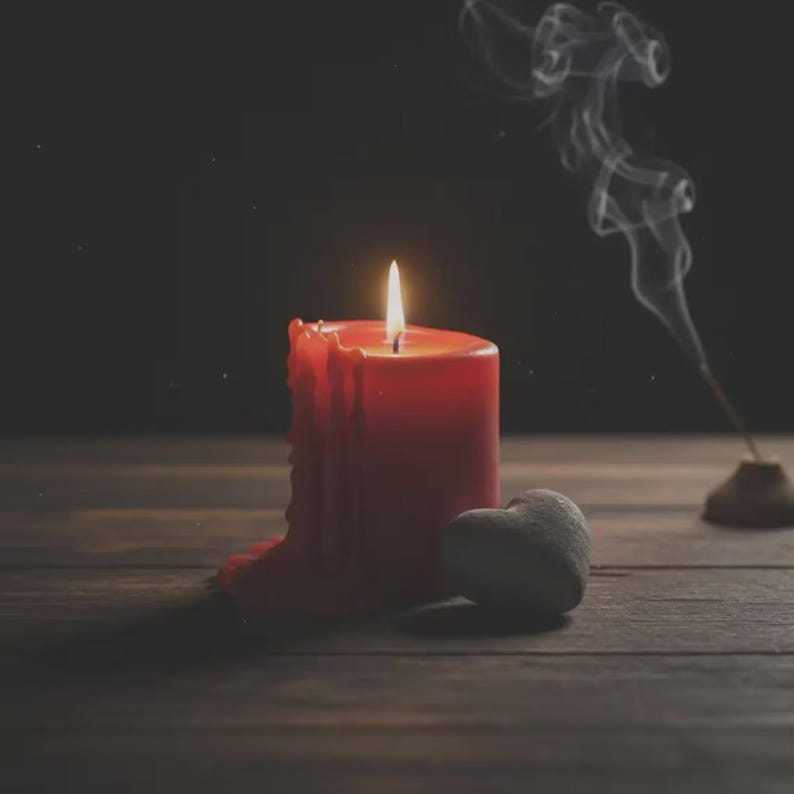 May include: A lit red candle with melted wax, a small heart-shaped stone, and a smoking incense stick on a wooden surface. The candle flame glows brightly against the dark background, creating a warm and inviting ambiance.