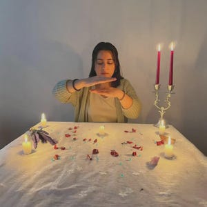 May include: A woman with her hands raised, positioned above a table covered with a white cloth. The table is adorned with lit candles, rose petals, and other decorative items. Two red candles are in a decorative holder.
