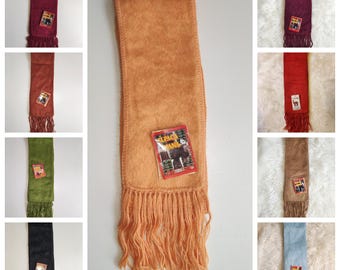 May include: A collection of alpaca wool scarves in various colors, including orange, burgundy, brown, green, black, red, and blue. Each scarf has a fringed edge and a tag with an alpaca image. The scarves are rectangular and appear soft.