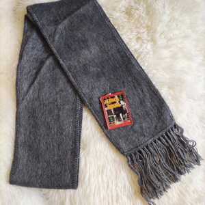 May include: A gray scarf with fringe at the ends. The scarf has a tag that reads "Alpaca Diana". The scarf is made of a soft, fuzzy material and is laying on a white, fluffy surface.