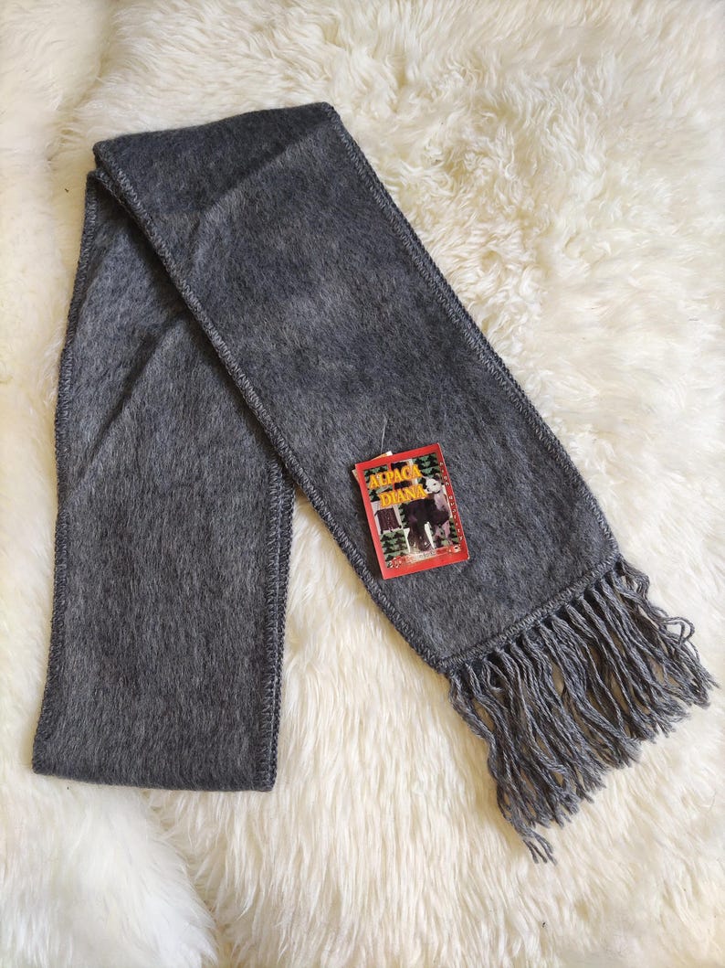 May include: A gray scarf with fringe at the ends. The scarf has a tag that reads "Alpaca Diana". The scarf is made of a soft, fuzzy material and is laying on a white, fluffy surface.