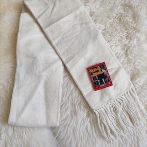 May include: Cream-colored scarf with fringe detail and a tag that reads "ALPACA DIANA". The scarf is made of a soft, textured material and is laid out on a fluffy, white surface. The scarf is a winter accessory.