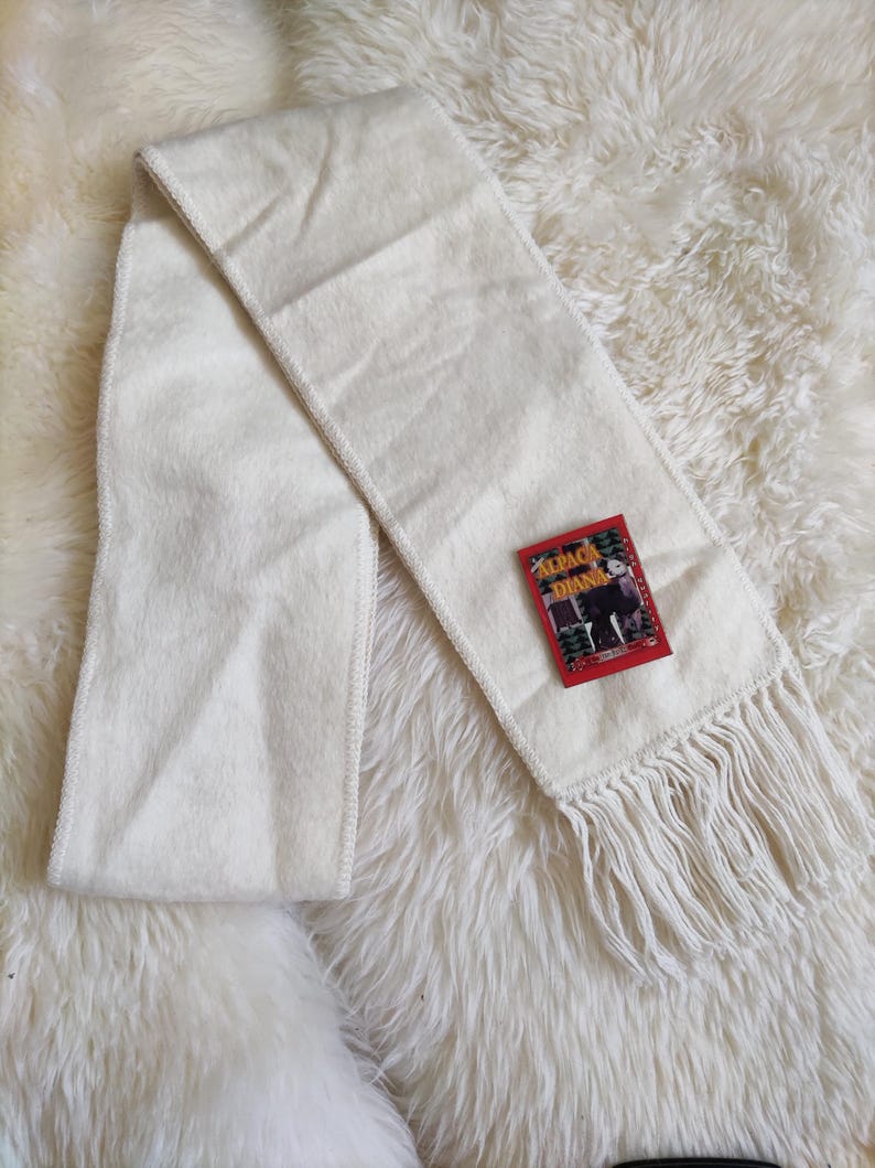 May include: Cream-colored scarf with fringe detail and a tag that reads "ALPACA DIANA". The scarf is made of a soft, textured material and is laid out on a fluffy, white surface. The scarf is a winter accessory.