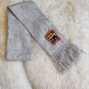 May include: A light gray scarf with fringe at the end and a small tag. The scarf is made of a soft, fuzzy material and is laid out on a white, fluffy surface. The tag has red, black, and white colors.