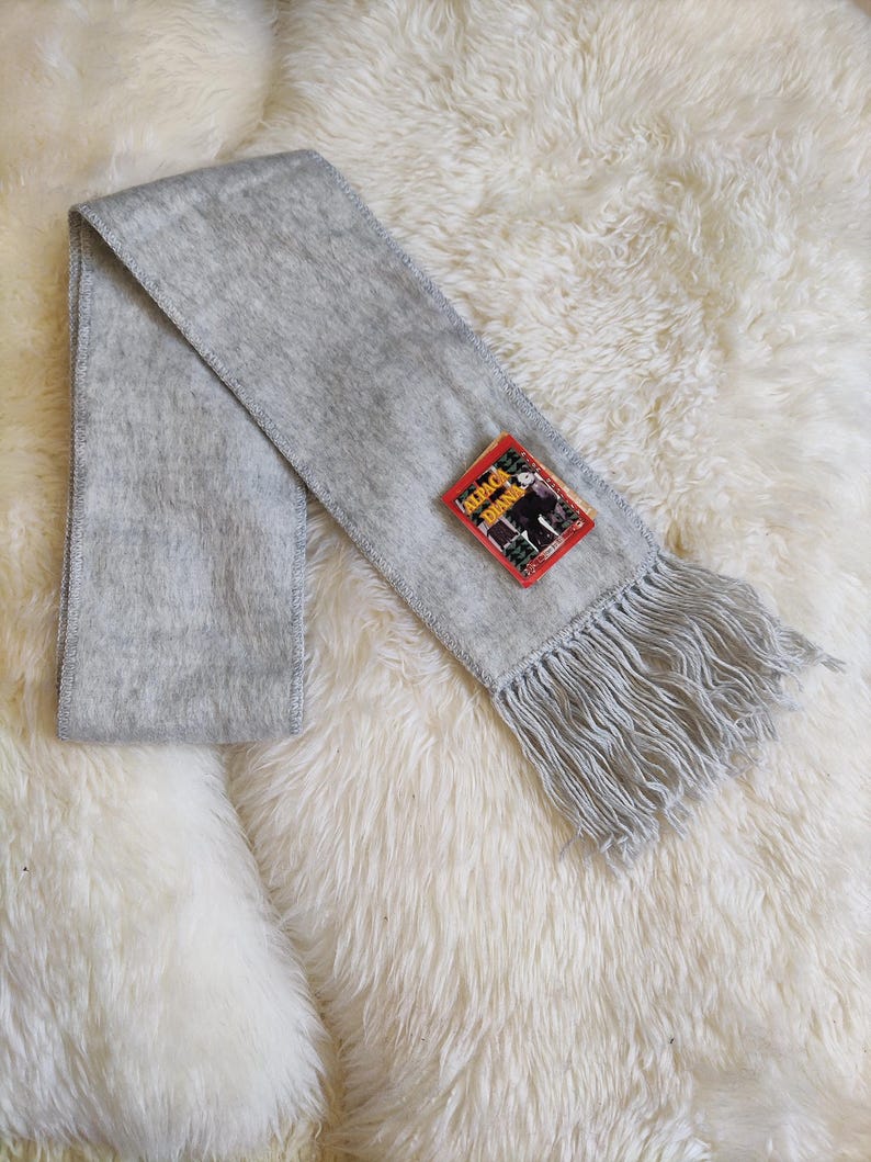 May include: A light gray scarf with fringe at the end and a small tag. The scarf is made of a soft, fuzzy material and is laid out on a white, fluffy surface. The tag has red, black, and white colors.
