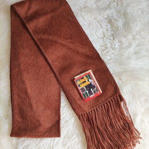 May include: A rust-colored scarf with a fringed edge and a small tag. The scarf is made of a soft, textured material and is laid out on a white, fluffy surface. The tag has text and an illustration.