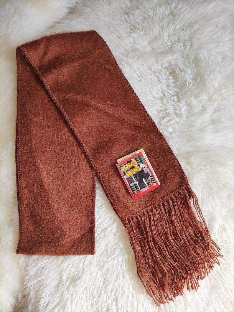 May include: A rust-colored scarf with a fringed edge and a small tag. The scarf is made of a soft, textured material and is laid out on a white, fluffy surface. The tag has text and an illustration.