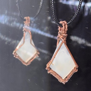 May include: Two handmade pendant necklaces. Each features a white, diamond-shaped stone encased in copper wire. The pendants hang from black cords. The background is dark and reflective, highlighting the jewelry's design.