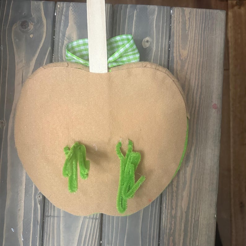 Puede incluir: Decoraci&oacute;n en forma de manzana de fieltro marr&oacute;n con detalles de limpiapipas verdes. Una cinta de cuadros verdes y blancos est&aacute; atada en la parte superior, sujeta a un palo de madera para colgar. La manzana tiene un contorno cosido.