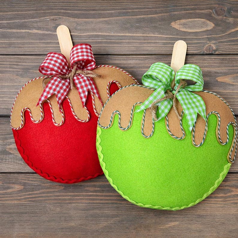 Puede incluir: Dos adornos de fieltro con forma de manzana, uno rojo y otro verde, con goteo de "caramelo" marr&oacute;n y un palito de madera. Cada manzana tiene un lazo de cuadros. Los adornos est&aacute;n sobre una superficie de madera.