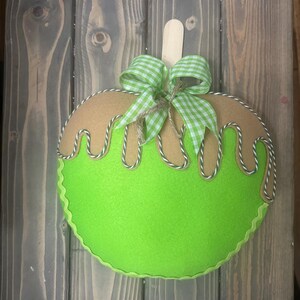 Puede incluir: Decoraci&oacute;n de fieltro con forma de manzana, con base verde claro y parte superior marr&oacute;n, que se asemeja a un helado. Cuenta con un lazo a cuadros verde y blanco, un palito de madera y ribetes blancos y verdes. La manzana mide unos 20 cm de di&aacute;metro.