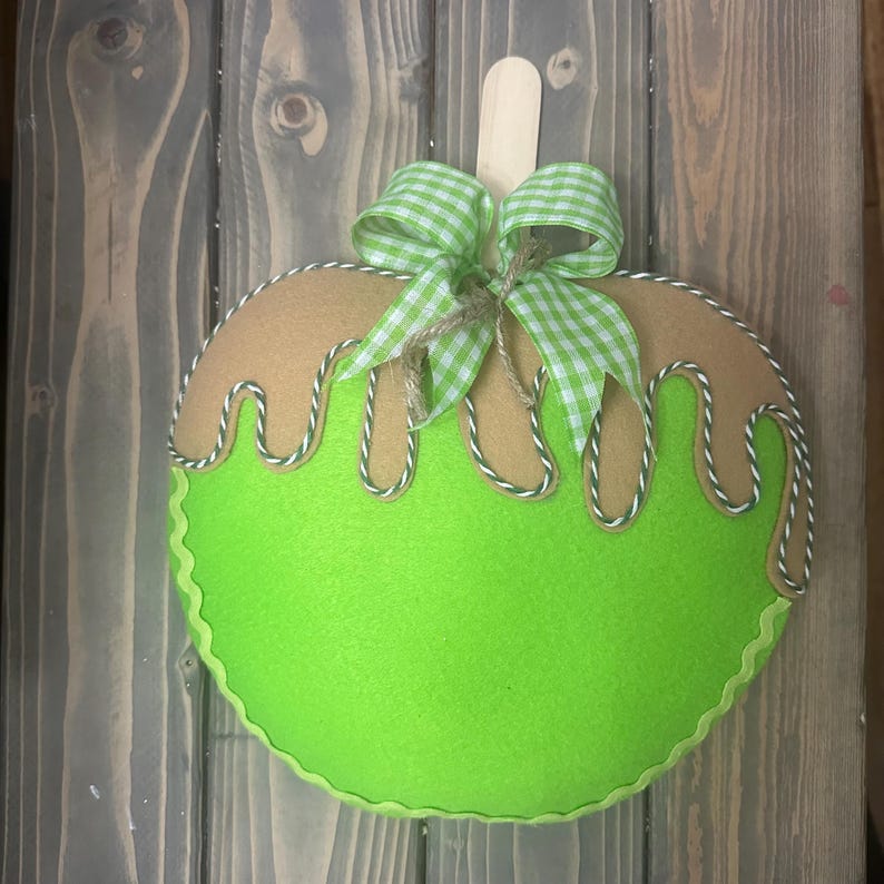 Puede incluir: Decoraci&oacute;n de fieltro con forma de manzana, con base verde claro y parte superior marr&oacute;n, que se asemeja a un helado. Cuenta con un lazo a cuadros verde y blanco, un palito de madera y ribetes blancos y verdes. La manzana mide unos 20 cm de di&aacute;metro.