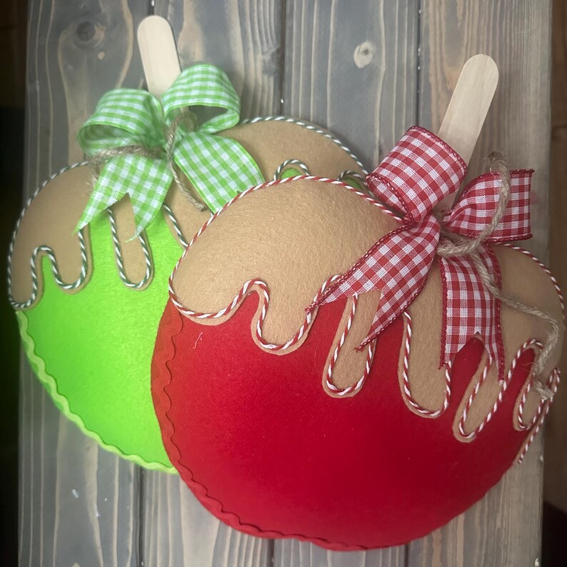 Puede incluir: Dos adornos de fieltro con forma de manzana con palitos de helado de madera. Uno es rojo con un lazo de cuadros rojos y blancos, el otro es verde con un lazo de cuadros verdes y blancos. Ambos tienen la parte superior de color beige y ribetes blancos y beige.