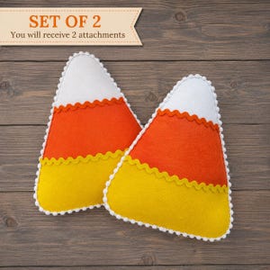 May include: Two felt candy corn decorations. Each candy corn is white, orange, and yellow, with a scalloped edge and white pom-pom trim. The text "SET OF 2" and "You will receive 2 attachments" is in the upper left corner.