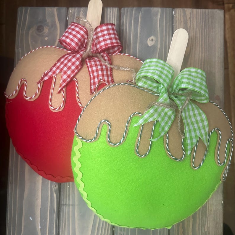 Puede incluir: Dos adornos de fieltro con forma de manzana, uno rojo y otro verde, con la parte superior de color canela y goteo blanco y marr&oacute;n. Cada uno tiene un palito de madera y un lazo de cuadros. La manzana roja tiene un lazo rojo y blanco, y la verde un lazo verde y blanco.