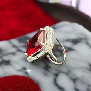 Peut inclure: Une bague en argent ornée d'une grosse pierre rouge rectangulaire facettée. La pierre est entourée d'un halo de petites pierres claires. La bague est posée sur une surface en marbre.