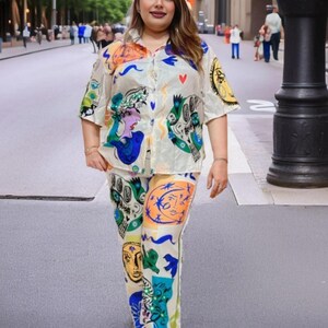 May include: A two-piece set featuring a short-sleeved button-down shirt and matching pants. The set is cream-colored and adorned with colorful abstract face and shape designs in blue, orange, green, and yellow. The outfit is worn outdoors.