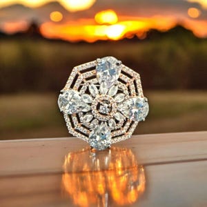 Peut inclure: Gros plan d'une bague étincelante au design géométrique. La bague présente un groupe central de pierres claires en forme de goutte d'eau, entouré de pierres rondes plus petites. Le sertissage est un mélange de tons or et argent, reflétant le coucher de soleil.