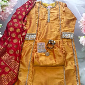 May include: Mustard yellow silk suit with silver trim and matching pants. A red and gold patterned scarf is draped on the side. Accessories include earrings, a bracelet, and a ring. The outfit is displayed on a white surface.
