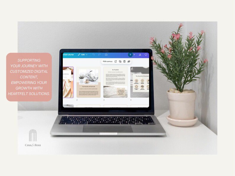May include: A laptop computer displaying a digital design program with four open pages. The pages feature a variety of designs, including a floral pattern, a beach scene, and a text-based page. The laptop is on a white table with a potted plant with pink flowers.