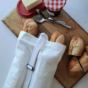 May include: A wooden cutting board with a loaf of bread in a white canvas bag, butter on a red dish, and a jar of jam. Two spoons and a knife are also on the board. Sliced bread is on the side.