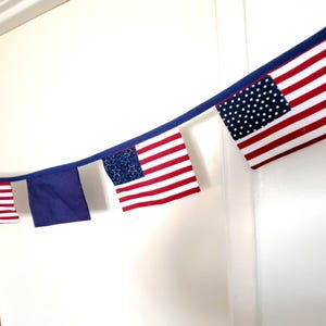 May include: A fabric bunting banner featuring a series of alternating pennants attached to a dark blue ribbon. The pennants include rectangular American flag designs with red and white stripes, blue fields with white stars, and solid blue squares.