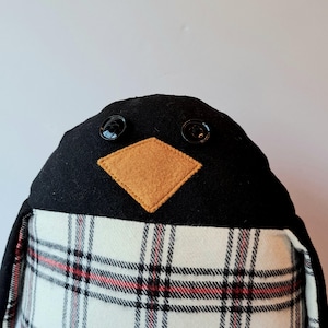 May include: A plush penguin with a black head, button eyes, and a tan diamond-shaped beak. The penguin's body is covered in a white and red plaid pattern. This soft toy is a charming decorative item.