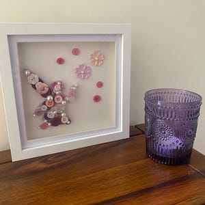 May include: A framed artwork featuring a purple and pink fairy made of buttons, with pink flowers and dots on a white background.
