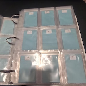 May include: A clear plastic trading card binder with multiple pockets, each containing a light blue card. Each card has a white sticker with a number. The binder is open, revealing the contents.