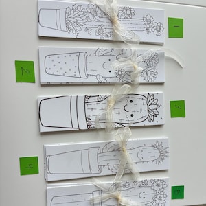 May include: Five white rectangular bookmarks with black line drawings of cacti in pots, tied with sheer ribbons. The cacti have smiling faces and floral accents. Green number stickers are visible.