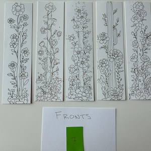 May include: Five white rectangular bookmarks with black floral line drawings. The designs include various flowers, leaves, and a cactus. A small white card with the word "FRONTS" and a green rectangle with the number "7" are also present.