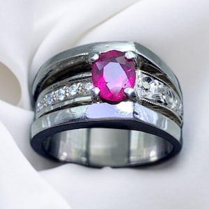 K&ouml;nnte beinhalten: Ein silberner Ring mit einem gro&szlig;en, ovalen Rubin-Edelstein, der von kleineren Diamanten umgeben ist. Der Ring hat ein breites Band mit einer schwarzen Einlage.