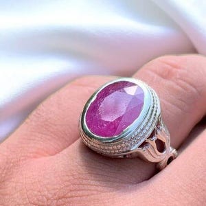 Op de afbeelding: Een zilveren ring met een grote, ovale, roze robijn die is gezet in een decoratieve zilveren band.