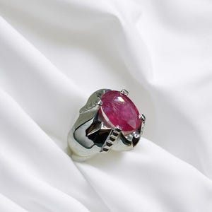 K&ouml;nnte beinhalten: Ein silberner Ring mit einem gro&szlig;en, ovalen, rosafarbenen Edelstein, der in einer Krappenfassung sitzt. Der Ring hat ein detailliertes Band mit einem strukturierten Design.